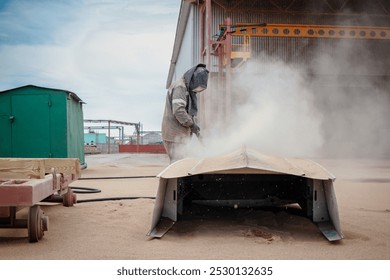 A worker in protective gear sandblasts a metal structure to eliminate rust at an industrial plant, creating clouds of dust in the daytime environment. - Powered by Shutterstock