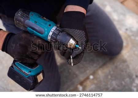 Worker in protective gloves inserting drill bit into cordless drill chuck, preparing power tool for drilling work.