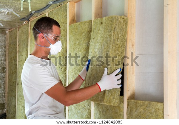 Worker in protective goggles and respirator\
insulating rock wool insulation in wooden frame for future house\
walls for cold barrier. Comfortable warm home, economy,\
construction and renovation\
concept