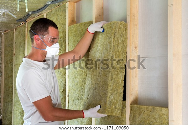 Worker in protective goggles and respirator\
insulating rock wool insulation in wooden frame for future house\
walls for cold barrier. Comfortable warm home, economy,\
construction and renovation\
concept