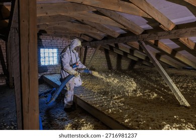 Worker in protective suit uses blue hose to spread loose fill insulation in a timber framed attic with exposed rafters and a glass block window under harsh side light.
