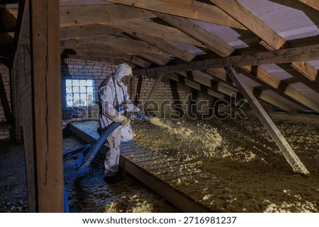 Worker in protective suit uses blue hose to spread loose fill insulation in a timber framed attic with exposed rafters and a glass block window under harsh side light.