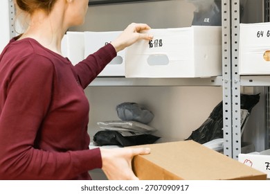 Worker pulling bin while holding package. Woman reaching for white numbered bin on shelf with brown box in hand
