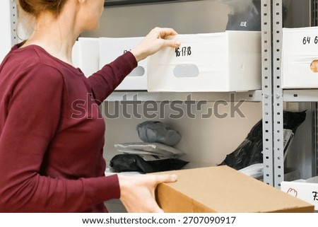 Worker pulling bin while holding package. Woman reaching for white numbered bin on shelf with brown box in hand