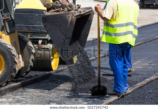 Worker put the hot asphalt\
on a street along tram car\'s railroad lines. Road construction\
workers with shovels in protective uniforms and an excavator. Hot\
summer day.