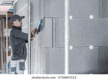 A worker puts insulation on a wall inside a building. He uses a tool to fit the material.