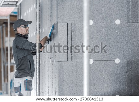 A worker puts insulation on a wall inside a building. He uses a tool to fit the material.