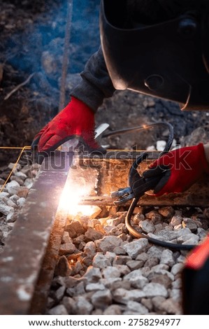 A worker in red gloves and a dark visor welds a steel rail joint on crushed ballast as sparks and smoke rise, with clamp, power leads, and alignment string in warm light.