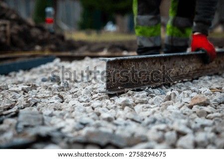 A worker in red gloves and high visibility pants drags a steel straightedge over crushed stone, daytime setting, shallow depth of field emphasizes precision and grit.
