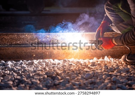 A worker in red gloves performs thermite welding on steel railroad tracks, sparks fly and blue white smoke rises over crushed ballast, ground level view at night.
