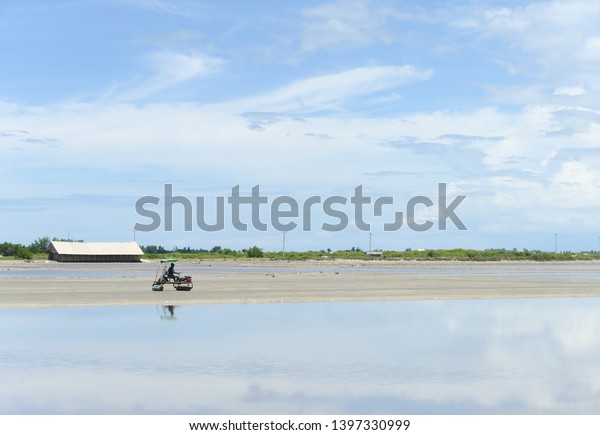 Worker\
riding compactor on dirt in salt farm near rural village on sunny\
day in summer with clear blue sky and clouds\
