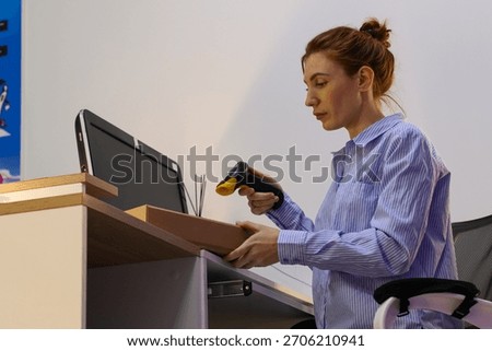 Worker scanning small package on desk at pickup point. Young woman in striped shirt holds box and uses barcode scanner at office desk with monitor.