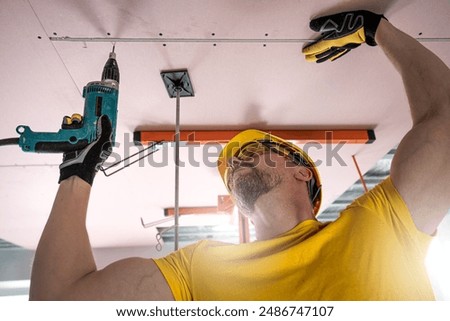 The worker screwing plasterboard to the ceiling. He is using special electric screwdriver and drywall lift. A red plasterboard improve the fire resistance of ceiling structures.