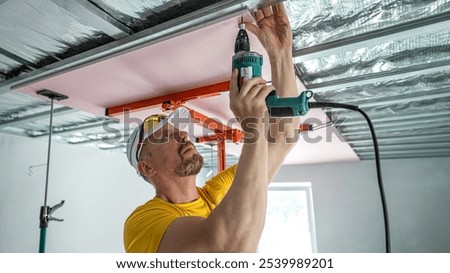 The worker screwing plasterboard to the ceiling. He is using special electric screwdriver and drywall lift. A red plasterboard improve the fire resistance of ceiling structures.