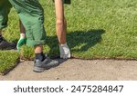 A worker is seen laying down a strip of sod in a garden. Their hands are visible, carefully placing the sod onto the ground.