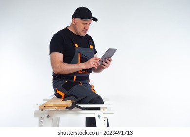 A Worker Sits On A Workbench And Looks At A Tablet Computer. Master With An Electronic Tablet In His Hands. The Carpenter Specifies The Details Of The Project Via The Internet.