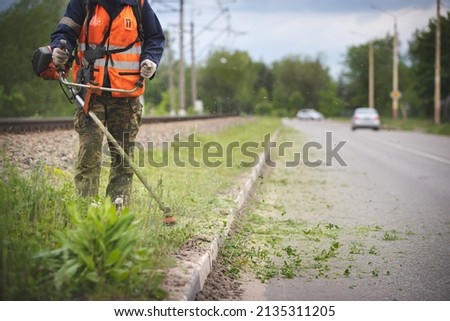 A worker in special reflective clothing with a gasoline mower in his hand. A man with a trimmer mows grass with dandelions on the side of the road