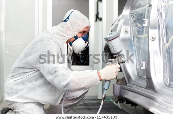 worker spraying grey paint on a car in special\
painting booth