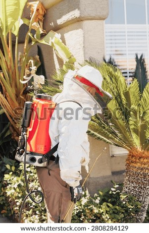 Worker sprays plants with red backpack sprayer. White hat and protective suit cover entire head. Sunlight glints off tall palms and stone wall. Fingers grip nozzle as person focuses ahead