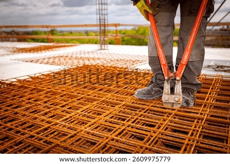 Worker is standing on pile of rusty rectangle steel, reinforcement for concrete as cutting reinforcement grid, mesh with bolt cutters at building site.