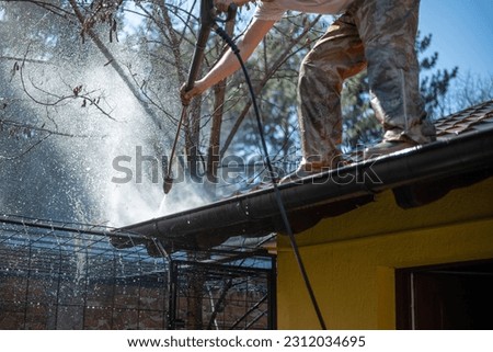 Worker standing on the roof and cleaning rain gutter with high pressure water jet. Professional using equipment for roof gutter cleaning. Maintenance and housekeeping concept of drainage channel. 