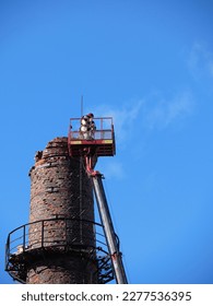 Worker stands on hydraulic lifting platform against slightly blurred background of old chimney. Industry. Construction equipment. Vertical photo.
