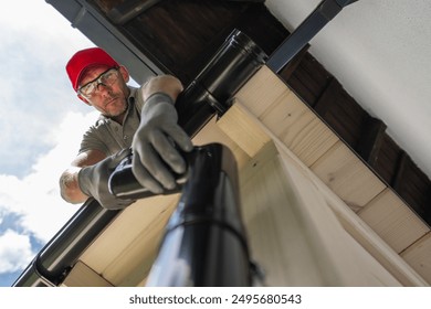 A worker stands on a roof, meticulously cleaning out gutters. Dressed in protective gear, he focuses on removing debris to ensure proper drainage. The bright sky adds to the atmosphere - Powered by Shutterstock