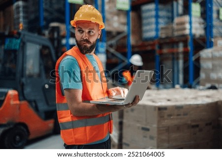 A worker stands in a warehouse inputting data on a laptop to manage inventory. Dressed in safety gear, he focuses on the screen, ensuring accurate record-keeping amidst shelves filled with supplies.