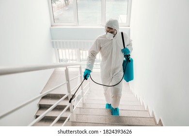 Worker in sterile uniform, with gloves and facial mask sterilizing railing in school.