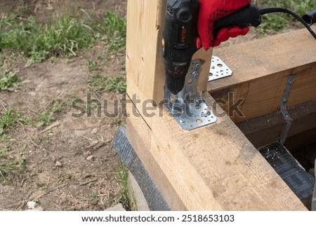 The worker strengthens the timber frame with the help of a tool and a metal angle. The craftsman tightens the screw in wooden beam while building a wooden house .