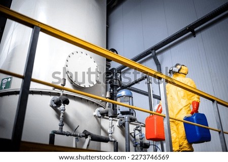 Worker supervisor in protective yellow suit, gas mask walking around large industrial acid tank and controlling production of chemicals.