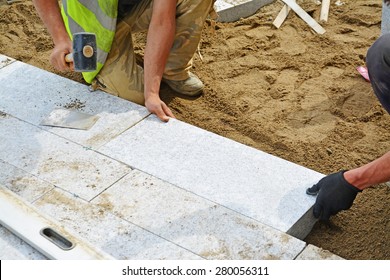 Worker Tapping Paver Into Place With Rubber Mallet. Installation Of Granite Paver Blocks Series With Motion Blur On Hammers And Hands.