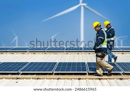 Worker Technicians are working to construct solar panels system on roof with wind turbine on background. Installing solar photovoltaic panel system. Renewable clean energy technology concept.