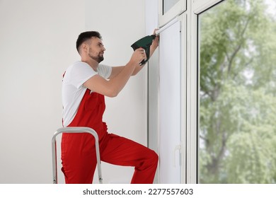 Worker in uniform installing roller window blind on stepladder indoors