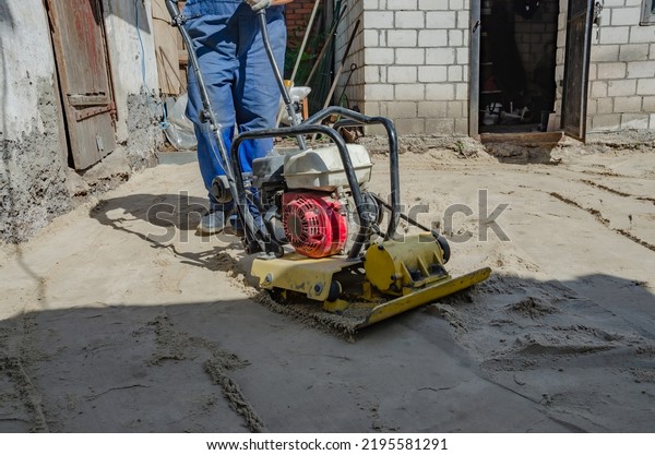 Worker in uniform and knee pads use vibratory plate\
compactor for path construction. Plate compactor for compaction\
soil or pavement or sidewalk. Indastrial equipment. Laying and\
tamping paving slabs