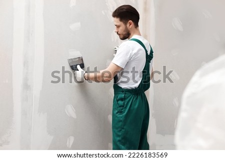 Worker in uniform plastering wall with putty knife indoors