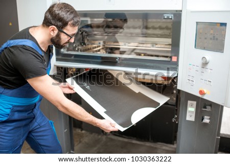 Worker in unifrom getting printed sheets from the offset machine at the printing manufacturing