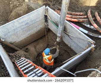 Worker uses hydro-vac method to safely expose buried gate valve inside trench box while electrical and telecom conduits remain live overhead.