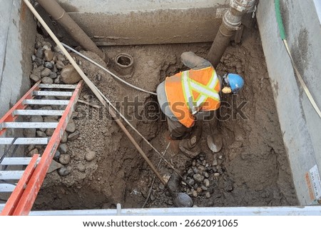 Worker uses hydro-vac method to safely expose buried gate valve inside trench box while electrical and telecom conduits remain live overhead.