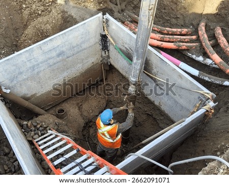 Worker uses hydro-vac method to safely expose buried gate valve inside trench box while electrical and telecom conduits remain live overhead.