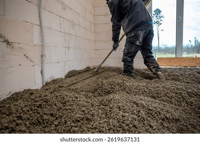 A worker uses a long handled tool to level wet concrete inside a building with white block walls. A large window reveals sparse trees and an open field.
