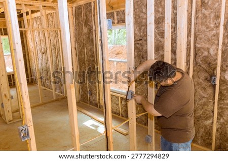 Worker uses measuring tool to mark wooden studs inside house progress in construction site.