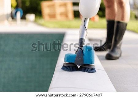 A worker uses a powered scrubbing machine to clean the deck of an empty residential swimming pool. The cleaning process focuses on outdoor maintenance and property upkeep.