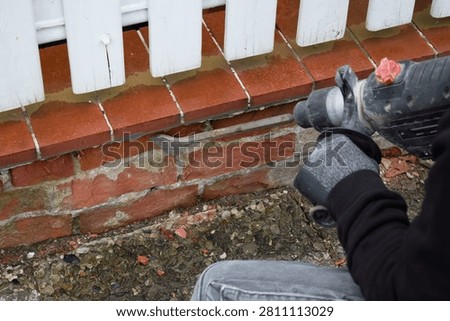 A worker uses a rotary hammer to remove the old cladding from a brick porch. The renovation process involves removing tiles and the cement layer during the reconstruction of the building's facade. 