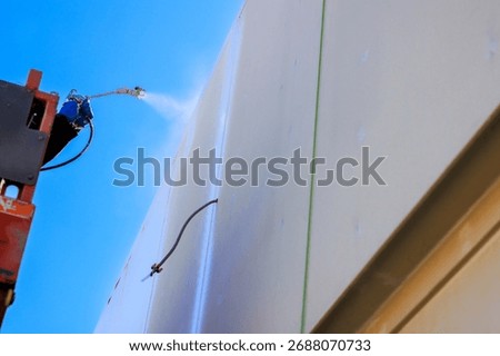 Worker uses spray tool apply paint on building wall while elevated on lifting equipment