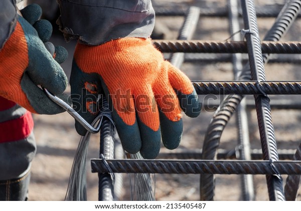 A worker uses steel tying wire to\
fasten steel rods to reinforcement bars. Close-up. Reinforced\
concrete structures - knitting of a metal reinforcing\
cage