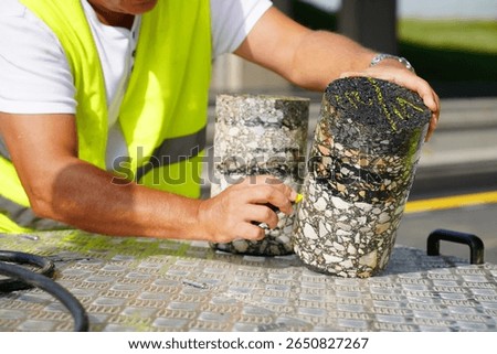 Worker Using Colored Chalk to Mark and Place Number on Cylindrical Core Sample from Asphalt Road	