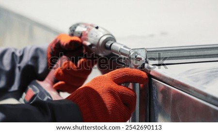 Worker Using Drill to Assemble Metal Frame. Close-up of a worker in orange gloves using a power drill to secure a metal frame with bolts.