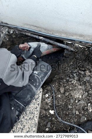 worker is using heavy hammer drill or jackhammer, excavate, drill into ground, masonry below ground level. work is taking place in close proximity to perimeter wall of house, installing new utilities, repairing waterproofing