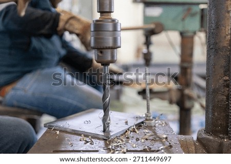 Worker using an industrial drill press to drill into a metal plate, showing precision in metalworking with safety gloves and equipment.
